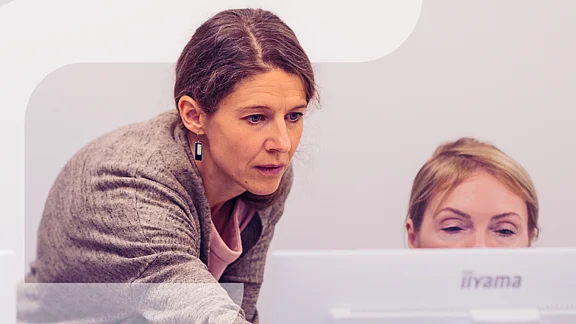 Zwei Frauen konzentrieren sich auf einen Computerbildschirm, eine steht und zeigt auf den Bildschirm, die andere sitzt davor. Im linken Bildbereich ist grafisch das stilisierte "F" des Projekts „Fempower“ eingeblendet.