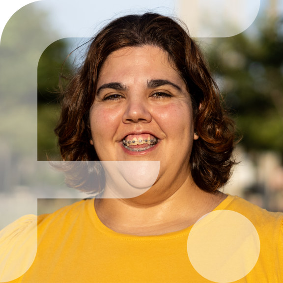 Eine fröhliche Person mit braunen, schulterlangen Haaren und Zahnspange lächelt in die Kamera. Sie trägt ein leuchtend gelbes Oberteil. Im Hintergrund sind unscharf Bäume und Gebäude zu erkennen. Im linken Bildbereich ist grafisch das stilisierte "F" des Projekts „Fempower“ eingeblendet.