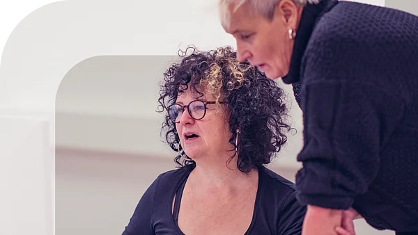 Zwei Frauen arbeiten gemeinsam am Computer, eine sitzt und zeigt auf den Bildschirm, die andere steht und schaut konzentriert zu. Im linken Bildbereich ist grafisch das stilisierte "F" des Projekts „Fempower“ eingeblendet.