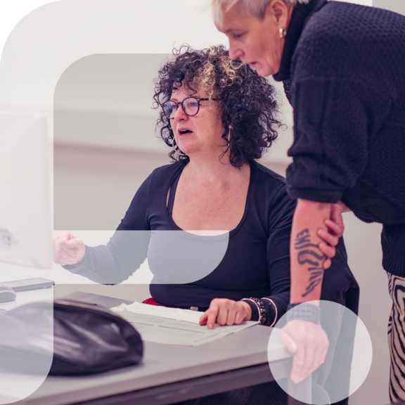 Zwei Frauen arbeiten gemeinsam am Computer, eine sitzt und zeigt auf den Bildschirm, die andere steht und schaut konzentriert zu. Im linken Bildbereich ist grafisch das stilisierte "F" des Projekts „Fempower“ eingeblendet.