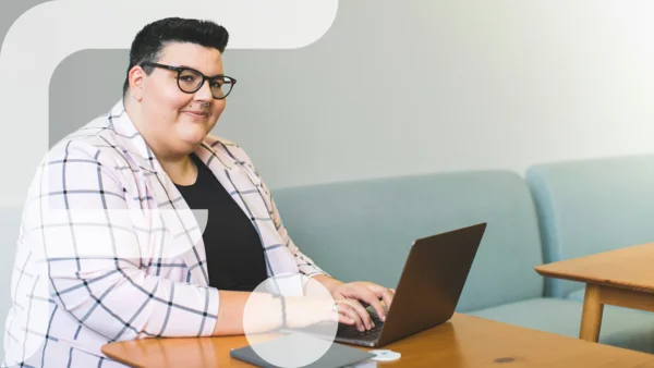 Eine selbstbewusst lächelnde Person mit Kurzhaarschnitt und Brille sitzt an einem Holztisch auf einem hellblauen Sofa und tippt auf einem Laptop. Sie trägt ein kariertes Sakko über einem schwarzen Shirt. Auf dem Tisch liegen ein Tablet und ein rundes weißes Gerät. Im linken Bildbereich ist grafisch das stilisierte "F" des Projekts „Fempower“ eingeblendet.
