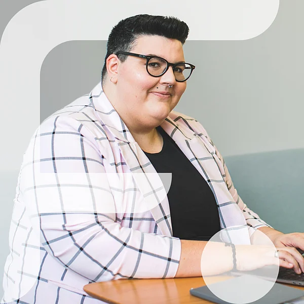Eine selbstbewusst lächelnde Person mit Kurzhaarschnitt und Brille sitzt an einem Holztisch auf einem hellblauen Sofa und tippt auf einem Laptop. Sie trägt ein kariertes Sakko über einem schwarzen Shirt. Auf dem Tisch liegen ein Tablet und ein rundes weißes Gerät. Im linken Bildbereich ist grafisch das stilisierte "F" des Projekts „Fempower“ eingeblendet.