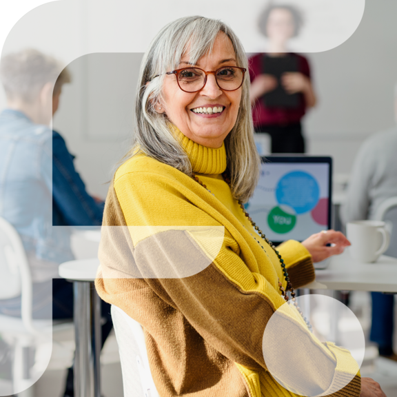 Eine ältere, lächelnde Person mit langen grauen Haaren und Brille sitzt in einem Seminarraum und blickt zur Kamera. Sie trägt einen gelb-braunen Pullover. Im Hintergrund sind andere Personen und ein Laptop mit einer bunten Präsentationsfolie zu sehen. Links im Bild ist grafisch das stilisierte „F“ des Projekts „Fempower“ eingeblendet.
